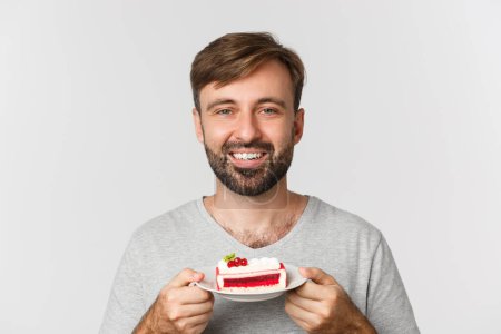 Foto de Primer plano de hombre sonriente guapo sosteniendo la torta, de pie sobre fondo blanco encantado - Imagen libre de derechos