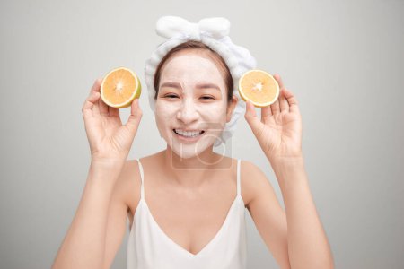 Téléchargez les photos : "Jeune asiatique ayant blanc masque d'argile faciale et tenant pièce orange." - en image libre de droit