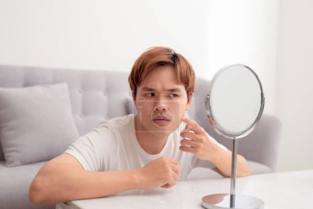 Téléchargez les photos : Bel homme concentré examinant de manière critique sa peau de visage dans le miroir et touchant ses joues avec les doigts - en image libre de droit