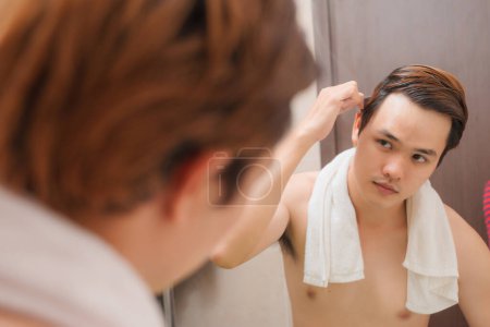 Photo for "Morning routine. Rear view of handsome young man combing his hair while standing against a mirror" - Royalty Free Image