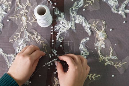 Téléchargez les photos : Gros plan des mains féminines de couturière tailleur couture robe de mariée en utilisant des perles et de la dentelle dans le studio - en image libre de droit