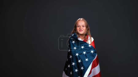 Téléchargez les photos : Petite fille patriote enveloppée dans un drapeau des États-Unis célèbre la fête de l'indépendance exprime patriotisme isolé sur fond noir - en image libre de droit