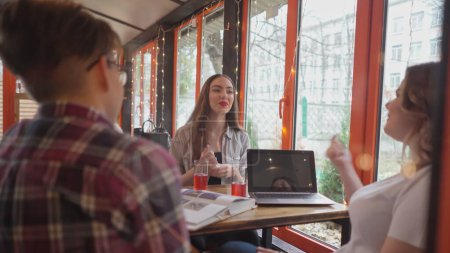Téléchargez les photos : Étudiants un gars et des filles parlant assis ensemble étudient dans un café avec des lumières en arrière-plan alors qu'ils s'assoient à une table avec des livres ouverts et un ordinateur portable dessus. Images 4K - en image libre de droit
