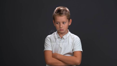 Téléchargez les photos : Petit garçon malheureux ou triste regardant un côté fronçant les sourcils portant un polo blanc et un pantalon noir isolé sur fond noir - en image libre de droit