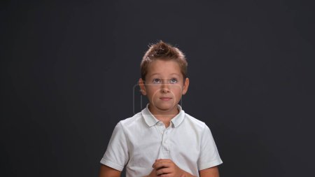 Photo for Expecting miracle boy in white shirt holding hands together in front of him while looking sideways at upper corner. Isolated on black background - Royalty Free Image