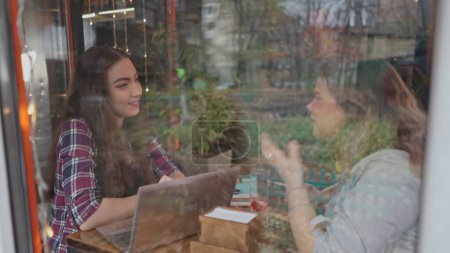 Foto de Ver a través de vidrio dos estudiantes chicas hablando mientras disfrutan del café juntos en una cafetería con los clientes caminando en el fondo mientras se sientan en una mesa charlando trabajando en el ordenador portátil - Imagen libre de derechos