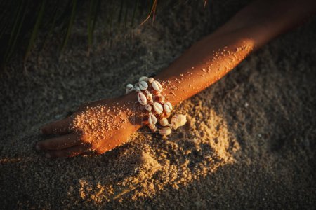 Foto de Tiro recortado de brazo femenino con brazalete de conchas marinas en la playa de arena - Imagen libre de derechos
