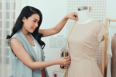 Foto de "Una chica asiática está trabajando en el taller. Ella hace ajuste en el vestido en el maniquí
." - Imagen libre de derechos