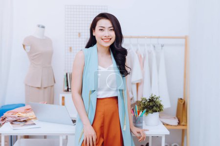 Téléchargez les photos : "Jeune styliste asiatique souriant et debout devant le bureau dans l'atelier
" - en image libre de droit