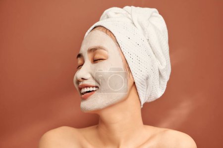 Téléchargez les photos : "Gros plan portrait d'une jolie fille avec une serviette sur la tête et un masque d'argile sur le visage isolé sur fond orange." - en image libre de droit