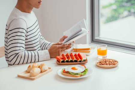Téléchargez les photos : Homme manger le petit déjeuner et lire le livre à la maison - en image libre de droit