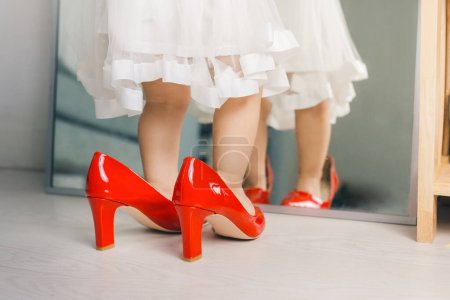 Téléchargez les photos : "Mignon drôle petite fille marche à la maison en essayant chaussures à talons hauts rouges de maman" - en image libre de droit