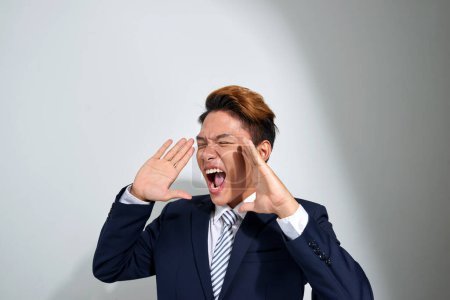 Foto de "joven asiático hombre de negocios gritando con las manos ventosas a su boca, aislado en gris fondo
" - Imagen libre de derechos