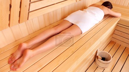 Téléchargez les photos : "Femme se relaxant dans un sauna en bois couché sur le ventre sur le banc avec une serviette enroulée autour de son corps" - en image libre de droit