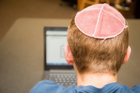 Photo for "Young boy wearing a yarmulke from the back doing work on a laptop in a library with colorful books on shelves." - Royalty Free Image