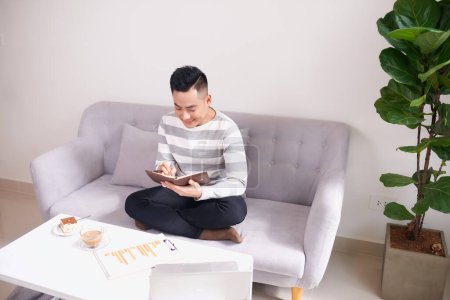 Téléchargez les photos : Jeune homme d'affaires heureux écrivant des notes sur le canapé et regardant ordinateur portable - en image libre de droit