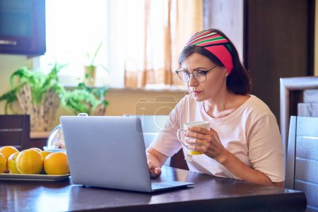 Téléchargez les photos : Femme d'âge moyen assise à la maison dans la cuisine avec un ordinateur portable et une tasse de café - en image libre de droit