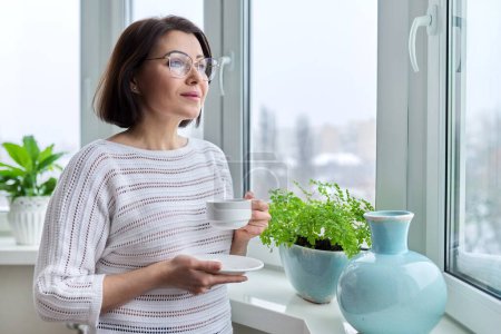 Téléchargez les photos : "Moyen âge belle femme avec des lunettes avec une tasse de café à la maison en hiver automne saison" - en image libre de droit