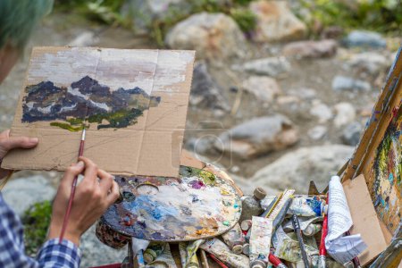 Photo for "Dombay, Russia 26 July 2020: Close up of young woman drawing picture on cardboard on hike. Paintress depicting mountain landscape outdoors." - Royalty Free Image