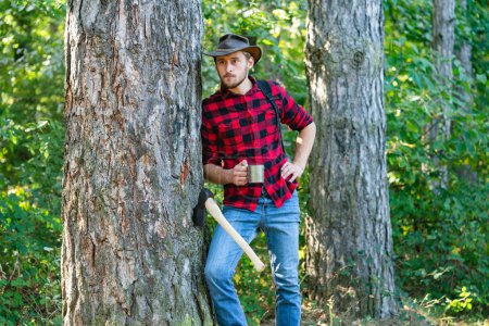 Foto de "Harvest of timber. Bearded man with axe concept. Deforestation. Handsome Woodworkers lumberjack plaid shirt holding the axe on green nature background." - Imagen libre de derechos