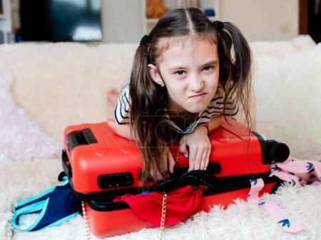 Téléchargez les photos : "petite fille appuyé sur le dessus de la valise en essayant de le fermer. Une valise pleine de choses ne ferme pas." - en image libre de droit