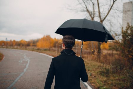 Téléchargez les photos : "Jeune homme en manteau noir marchant sous la pluie avec parapluie
" - en image libre de droit