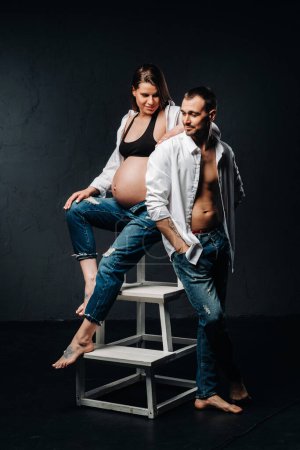 Téléchargez les photos : "une femme enceinte et un homme en chemise blanche et un jean dans un studio sur fond noir" - en image libre de droit