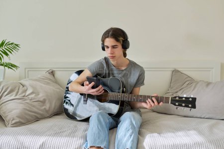 Photo for "Male teenager in headphones sitting at home with acoustic guitar and smartphone" - Royalty Free Image