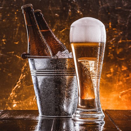 Téléchargez les photos : "deux bouteilles de bière dans un seau avec de la glace et un verre de bière avec une mousse luxuriante à côté d'un fond sombre" - en image libre de droit