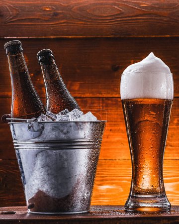 Téléchargez les photos : "deux bouteilles de bière dans un seau avec de la glace et un verre de bière avec une mousse luxuriante à côté d'un fond sombre" - en image libre de droit
