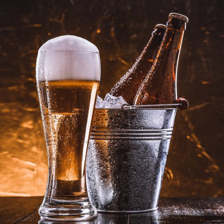 Foto de Dos botellas de cerveza en un cubo con hielo y un vaso de cerveza con espuma exuberante junto a un fondo oscuro - Imagen libre de derechos