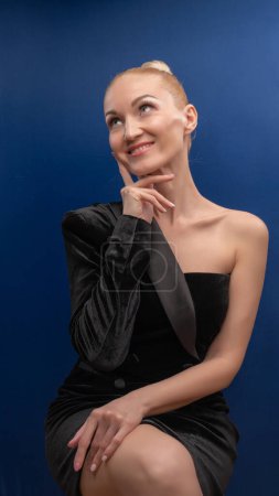 Foto de Buena chica madura se sienta en una silla, encantadora en un traje negro con un fondo de la cara europea en la luz del día blanco púrpura. Maquillaje. - Imagen libre de derechos