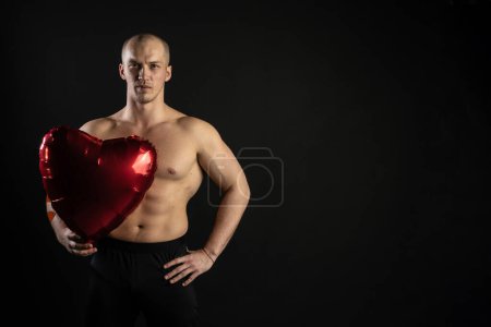 Téléchargez les photos : "Homme athlétique tenant un cœur rouge, un ballon nu dans sa main pour une journée de velentin pompé, bodybuilder romantique. amant LGBT. Dans le studio sur un fond noir regarde joyeusement la caméra" - en image libre de droit