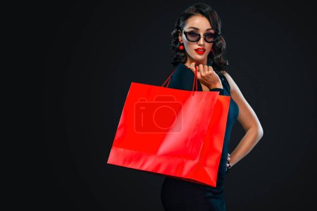 Photo for Black Friday sale concept for shops. Shopping woman in sunglasses holding red bag isolated on dark background. - Royalty Free Image