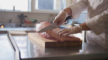 Photo for "Butcher cutting large piece of fresh raw meat lying on a wooden board in a commercial kitchen" - Royalty Free Image