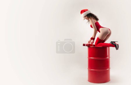 Photo for "A woman in a red Santa hat with Christmas gifts on a red barrel and a white background" - Royalty Free Image