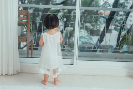 Téléchargez les photos : "Petite fille mignonne en belle robe est assis près de la fenêtre à la maison
." - en image libre de droit