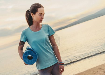 Téléchargez les photos : Femme avec tapis de yoga debout sur la plage - en image libre de droit