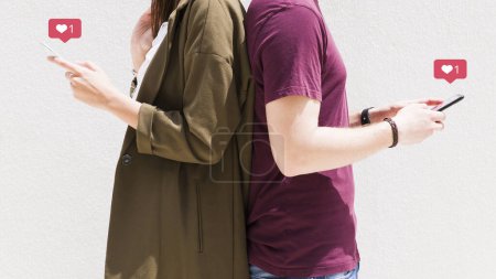 Téléchargez les photos : Couple debout en arrière en utilisant un téléphone portable avec des messages d'amour icônes contre mur - en image libre de droit