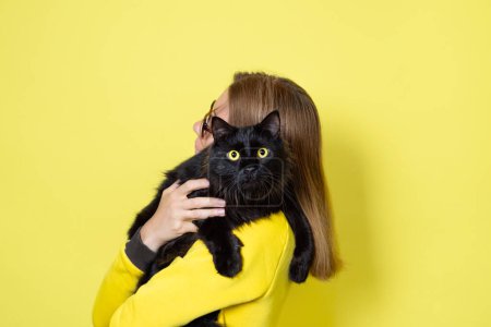 Téléchargez les photos : Fille dans une robe jaune tient son chat noir pelucheux bien-aimé avec des yeux jaunes sur un fond jaune. Concept de soins pour animaux - en image libre de droit