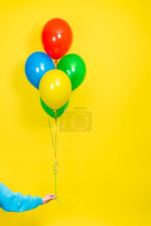 Foto de La mano femenina sostiene un montón de globos de colores. Parte o concepto actual. Globos verdes, rojos, amarillos y azules y mano aislada sobre fondo amarillo - Imagen libre de derechos