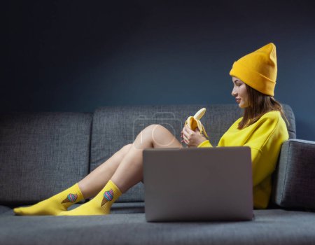 Téléchargez les photos : Femme sur canapé avec ordinateur portable manger banane sexy - en image libre de droit