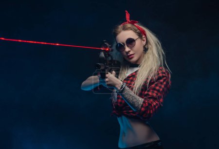 Téléchargez les photos : Une blonde lumineuse dans un chapeau rouge avec une mitrailleuse sur un fond sombre - en image libre de droit