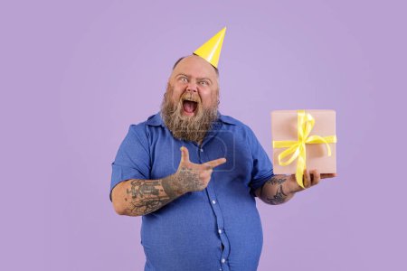 Téléchargez les photos : Homme excité avec surcharge pondérale dans des points de chemise serrés sur boîte-cadeau sur fond violet - en image libre de droit