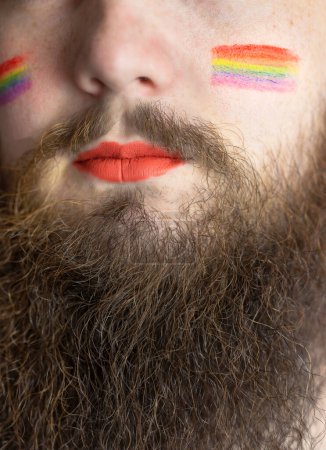 Téléchargez les photos : Jeune homme barbu beau avec le drapeau de fierté sur sa joue, drapeau arc-en-ciel debout pour LGBTQ, Droit de genre et minorité sexuelle. Portrait - en image libre de droit