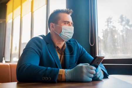 Photo for "Caucasian male businessman in protective gloves and medical mask spends time in mobile phone in cafe" - Royalty Free Image