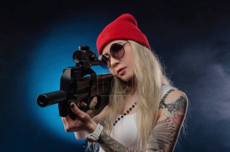 Téléchargez les photos : Blonde lumineuse dans un chapeau rouge avec une mitrailleuse sur un fond sombre - en image libre de droit