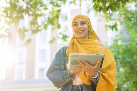 Téléchargez les photos : Une étudiante arabe. Belle étudiante musulmane portant un hijab jaune vif tenant une tablette
. - en image libre de droit