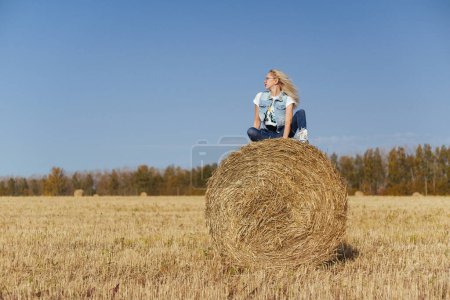 Téléchargez les photos : Belle villageoise posant en jeans sur une balle de foin dans un champ - en image libre de droit