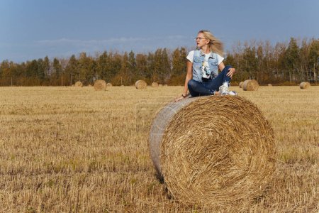 Photo for Beautiful girl villager posing in jeans on a bale of hay in a field - Royalty Free Image
